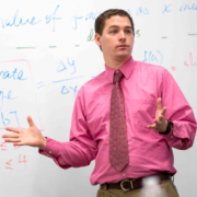 Clean-shaven white male with short dark hair wearing a pink oxford and tie standing in front of a whiteboard.