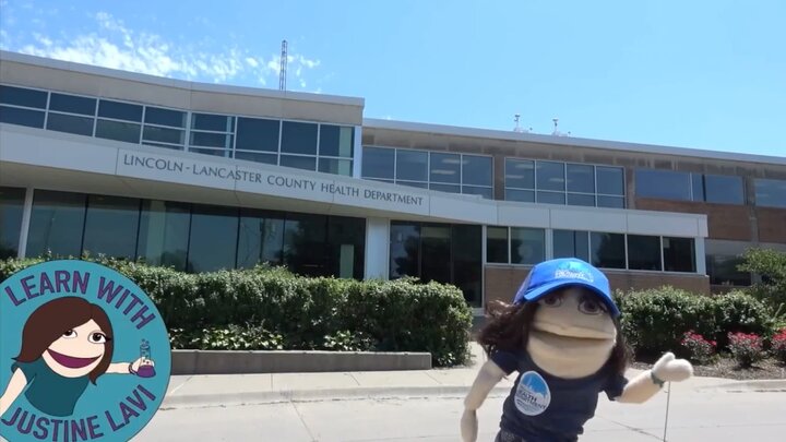 Screen video capture of Justine Lavi puppet in front of health department building