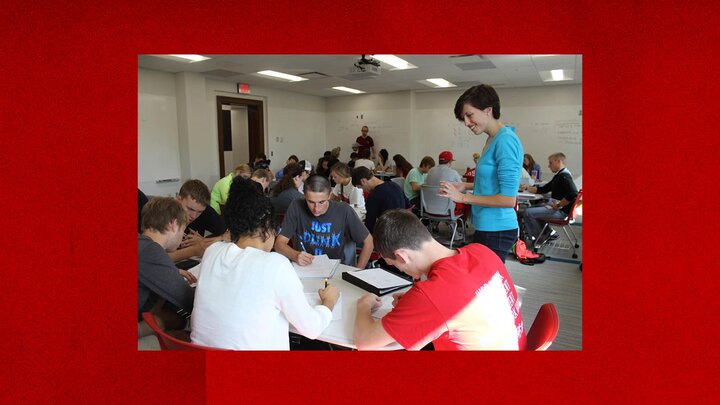 A classroom with students working around tables. In the foreground, several students are writing in notebooks while a facilitator stands to the right, smiling and engaging with the students.