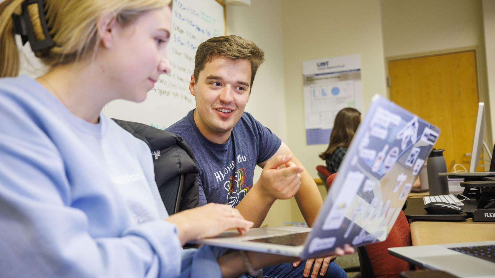 A smiling student points to a screen while talking with another student.